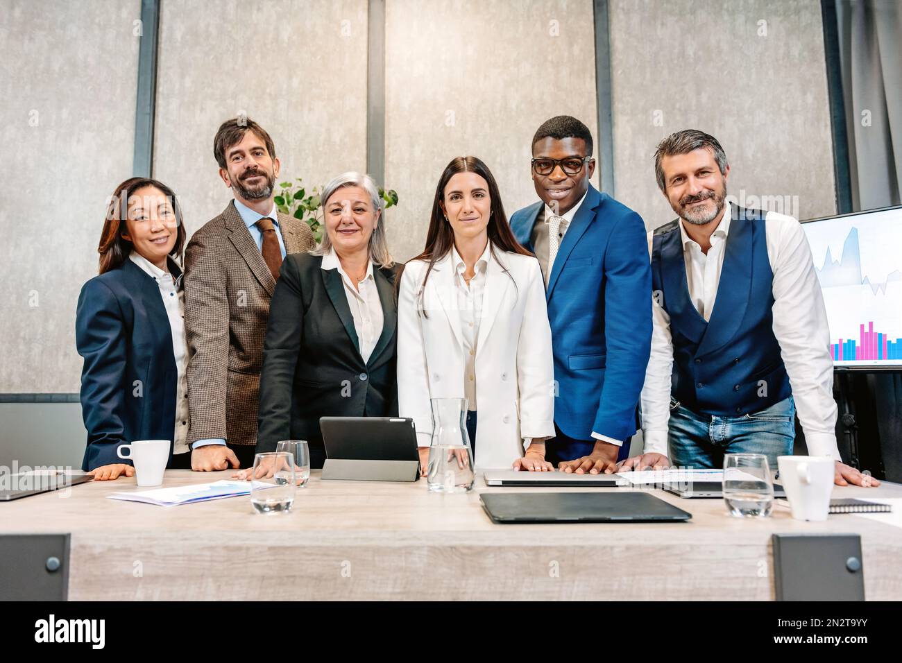Gruppo di colleghi multirazziali in abiti formali sorridenti e guardando la fotocamera mentre si è in piedi dietro il tavolo in una sala consiliare contemporanea Foto Stock