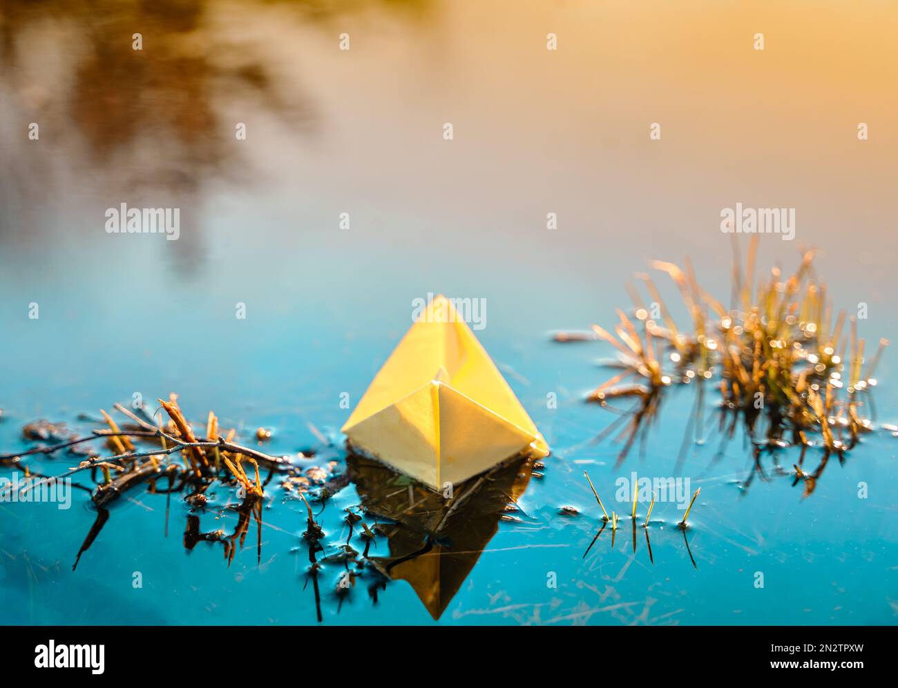 Imbarcazione di carta galleggiante gialla. Nave a vela colorata in grande pozza di primavera blu, acqua del fiume su strada invernale. Caldo tempo piovoso bagnato, erba vecchia. Ciao sp Foto Stock