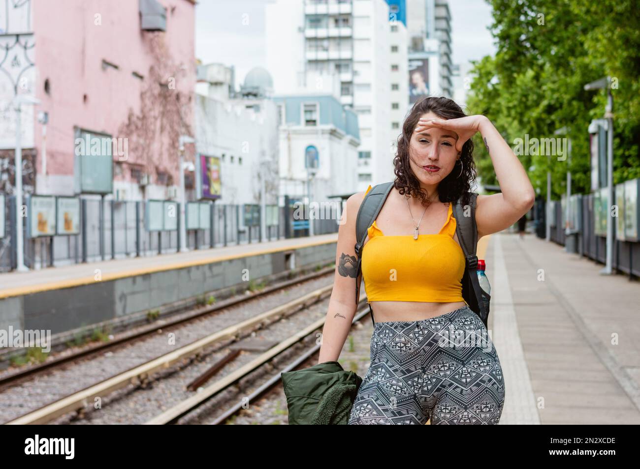 giovane donna latina di etnia argentina, con abiti gialli e uno zaino, è angosciata in piedi sulla piattaforma con la mano sulla fronte Foto Stock