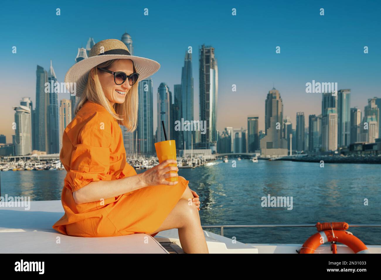 Donna sorridente in abito arancione con drink su uno yacht di lusso a Dubai al tramonto. skyline della città sullo sfondo. spazio di copia Foto Stock