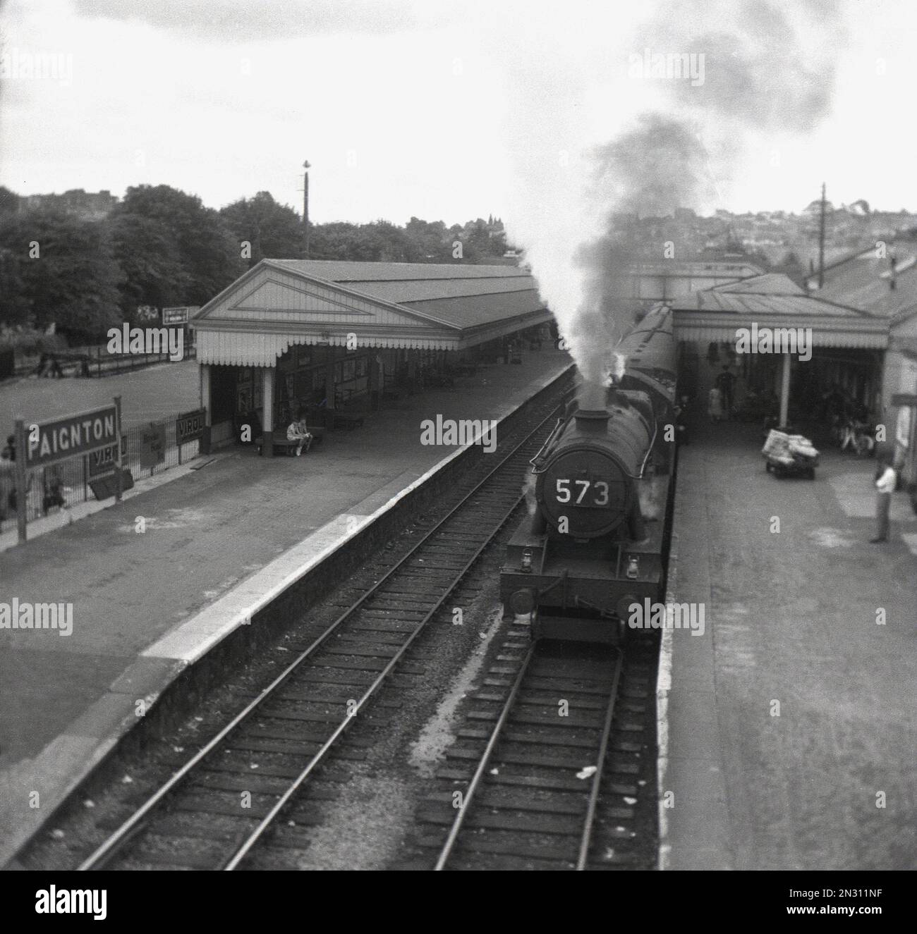 1955, storico, una locomotiva a vapore (573) in attesa sul binario alla stazione ferroviaria di Paignton, Torbay, Devon, Inghilterra, Regno Unito. Aperto come ferrovia a binario singolo nel 1859, costruito utilizzando un ampio calibro, nel 1892 è stato convertito in standard guage. La doppia pista arrivò nel 1910 e nel 1924 furono erette delle tettoie sulle piattaforme. Il treno sul paltform è un treno di classe Great Western 4700, una locomotiva a vapore utilizzata principalmente per i veloci servizi di trasporto notturno, ma è stato utilizzato anche per i servizi passeggeri verso l'Inghilterra occidentale durante i mesi estivi. Foto Stock