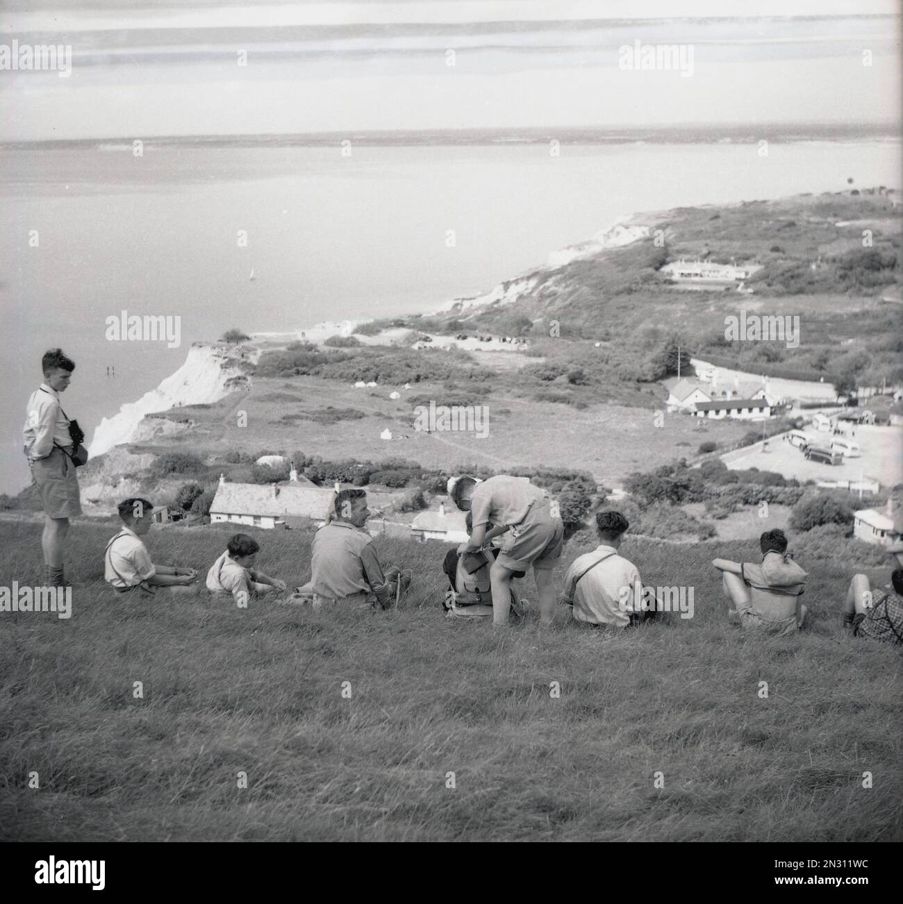 1955, storico, un insegnante di sesso maschile con un piccolo gruppo di ragazzi adolescenti allievi in un viaggio sul campo seduto sul pendio erboso che domina Alum Bay sull'Isola di Wight, Hampshire, Inghilterra, Regno Unito. Foto Stock