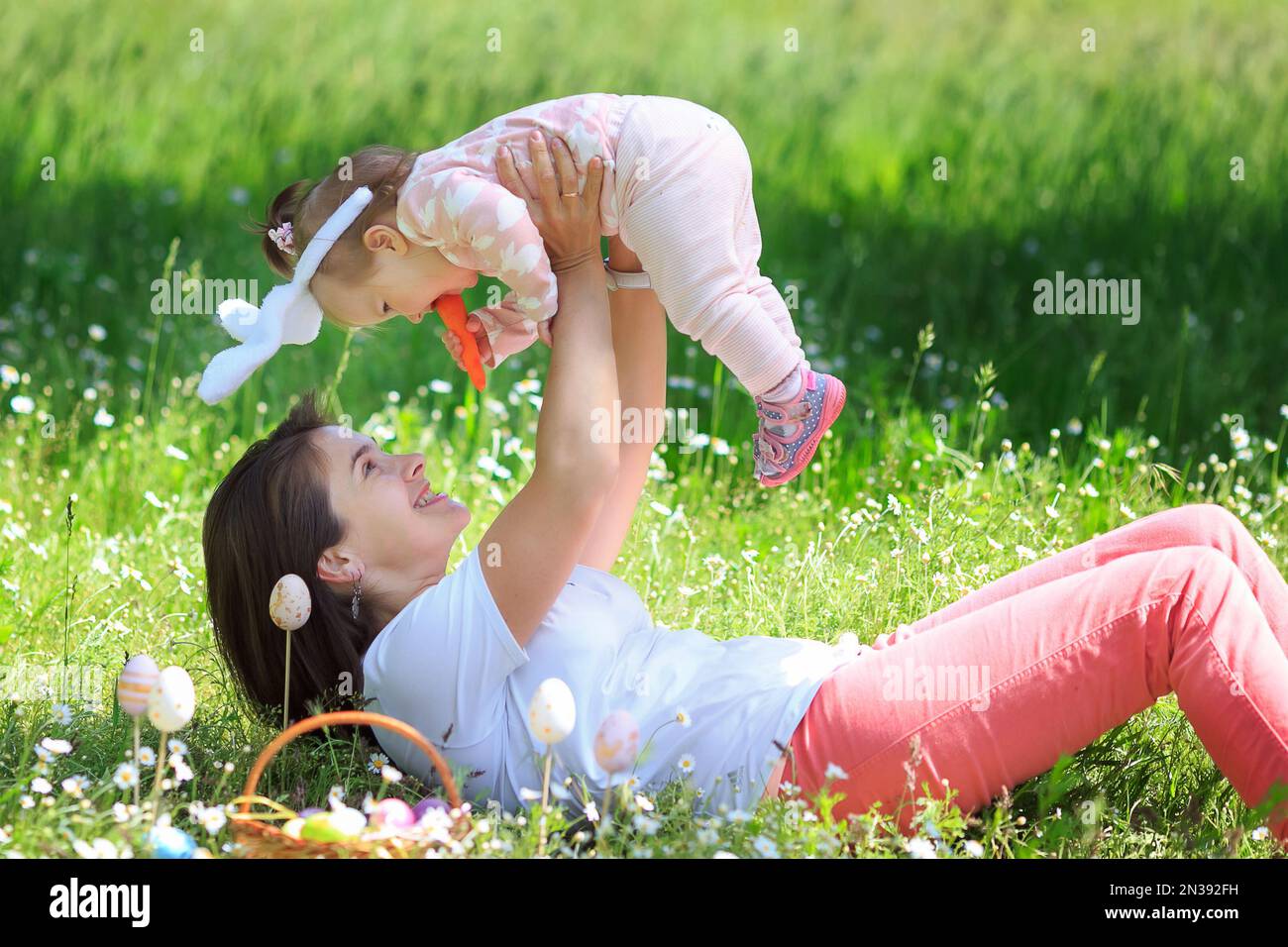 Mamma alza getta la figlia del bambino sul prato, divertendosi. Ragazza di un anno che gioca con la donna. Bambino piccolo, mamma che celebra la Pasqua all'aperto in camom Foto Stock