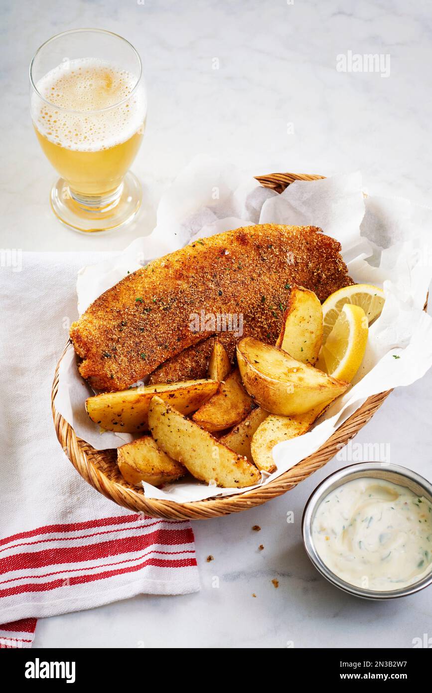 Tilapia impanata con spicchi di patate in un cestino, salsa tarter e un bicchiere di birra Foto Stock