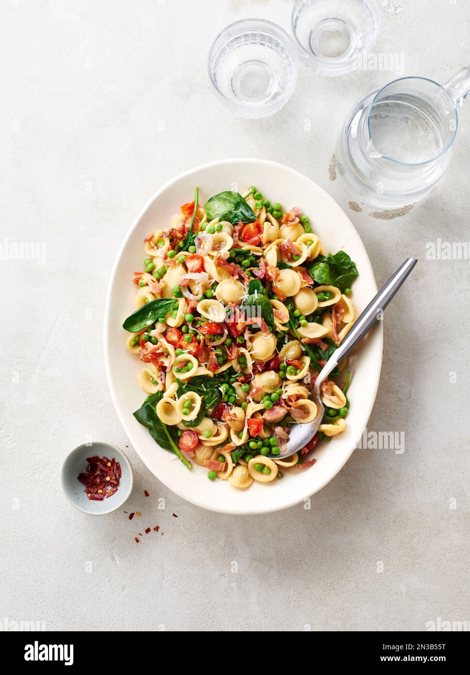 Piatto di orecchiette con piselli, pancetta, spinaci e pomodori con fiocchi di peperoncino in un recipiente piccolo, bicchieri d'acqua e caraffa sul dorso beige... Foto Stock