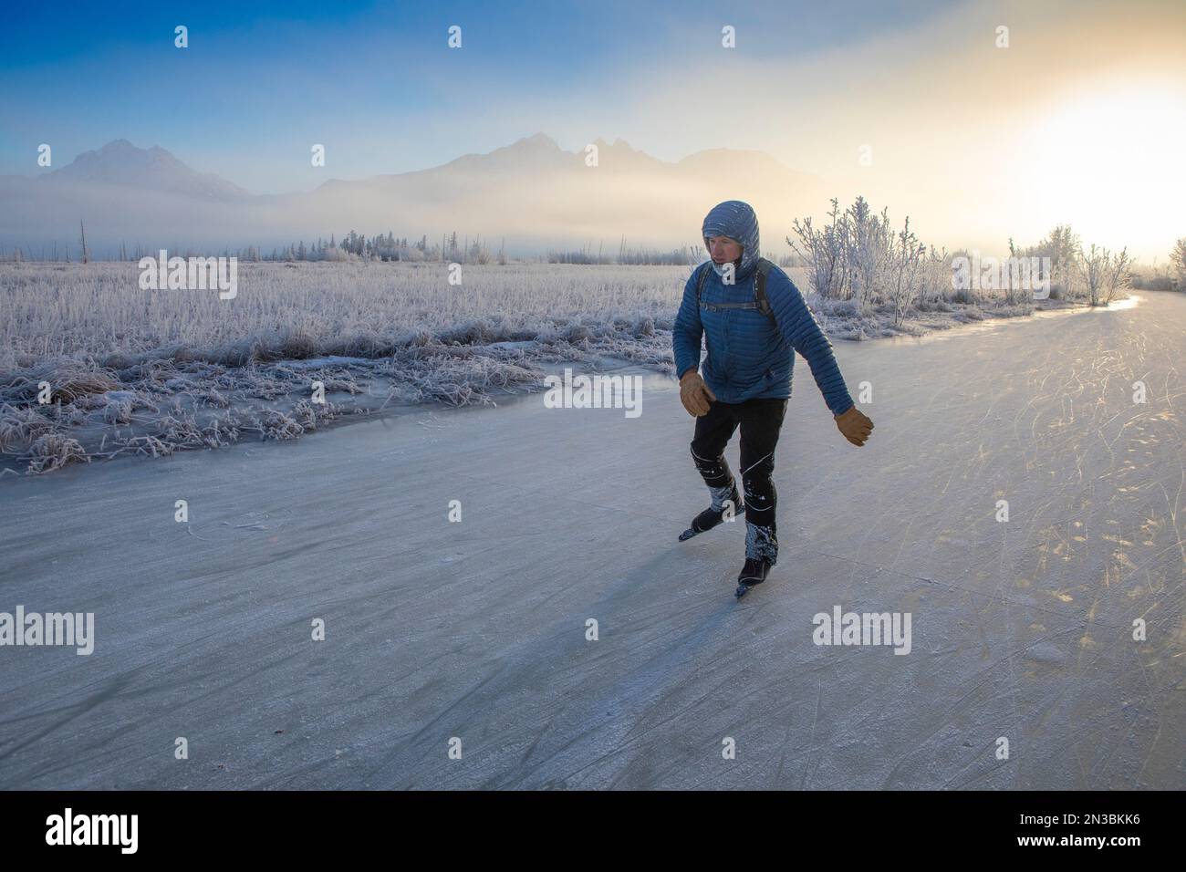 Un uomo caucasico pattinaggio su ghiaccio, pattinaggio nordico, lontano dal sole invernale in una giornata invernale parzialmente soleggiata, parzialmente nebbiosa, con ghiaccio coperto... Foto Stock