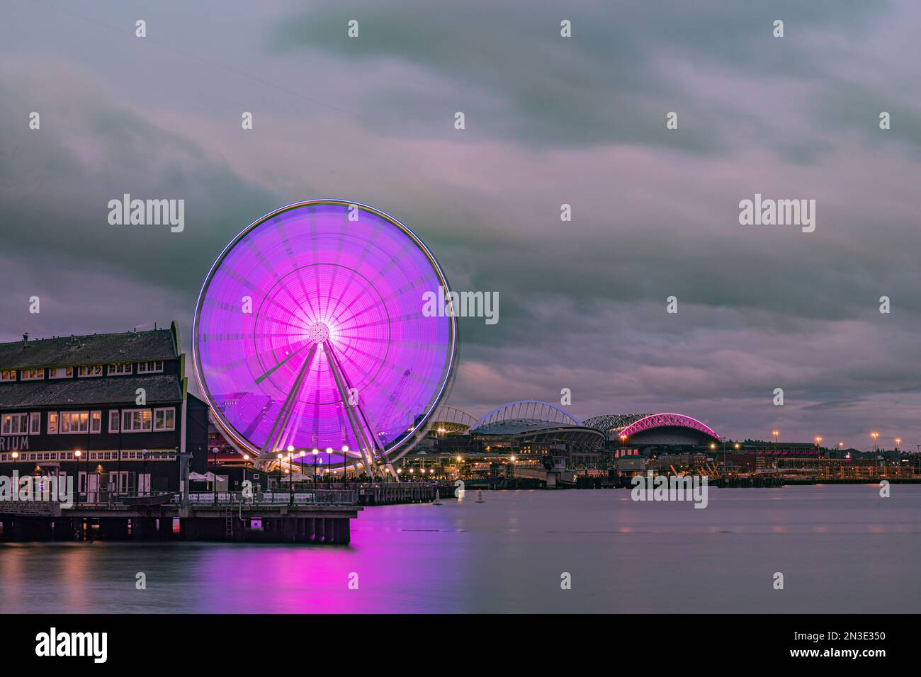 Una serata nuvolosa sul lungomare di Seattle guardando la Seattle Great Wheel che si riflette su Elliott Bay con Lumen Field e T-Mobile Park illuminati ... Foto Stock