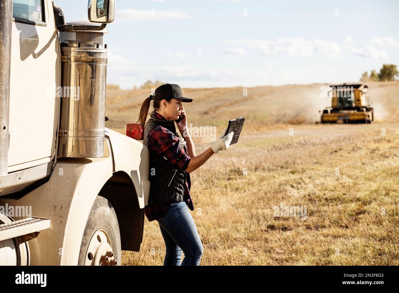Una donna in piedi accanto a un trasportatore di cereali, che utilizza dispositivi wireless portatili per gestire e monitorare un raccolto di Canola autunnale, che parla al telefono, con un... Foto Stock
