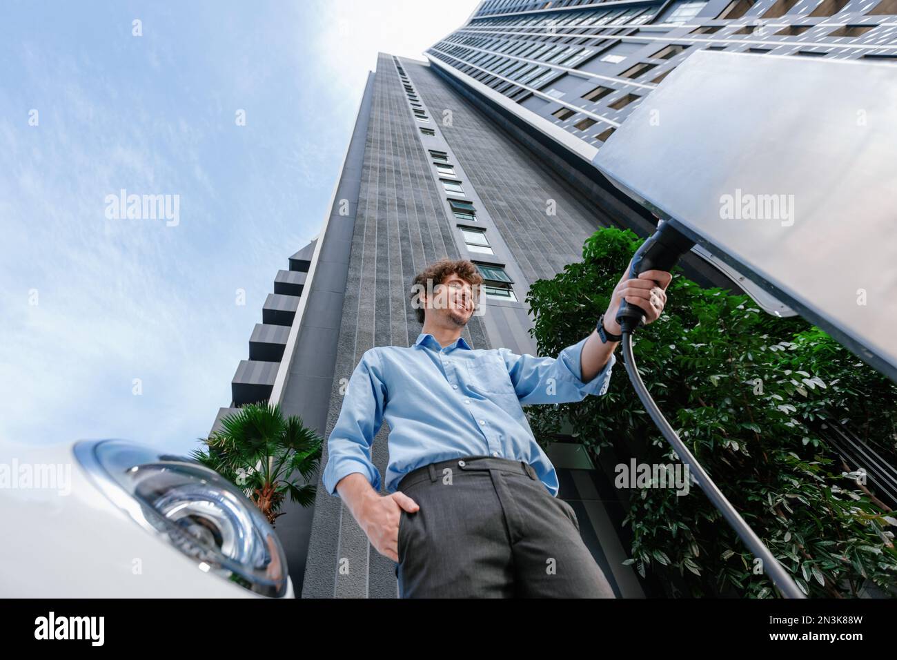 Fisheye vista vestito-rivestito uomo d'affari progressivo alla stazione di ricarica per veicolo elettrico nella città moderna con l'edificio residenziale condominio appartamenti Foto Stock