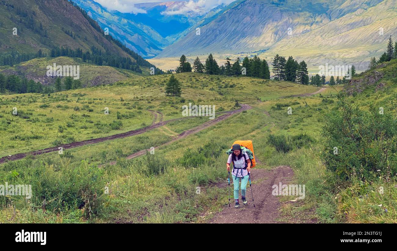 Una ragazza turistica con un grande zaino arancione e bastoni da trekking cammina wearily lungo la strada nel campo in estate vicino alle montagne e nuvole o Foto Stock