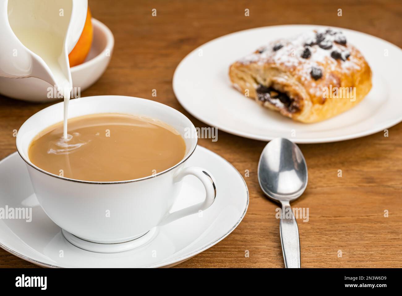 Versare il latte fresco nel caffè in una tazza di ceramica bianca su un piattino di ceramica con pasticceria danese ripiena di crema di cioccolato con gocce di cioccolato e. Foto Stock