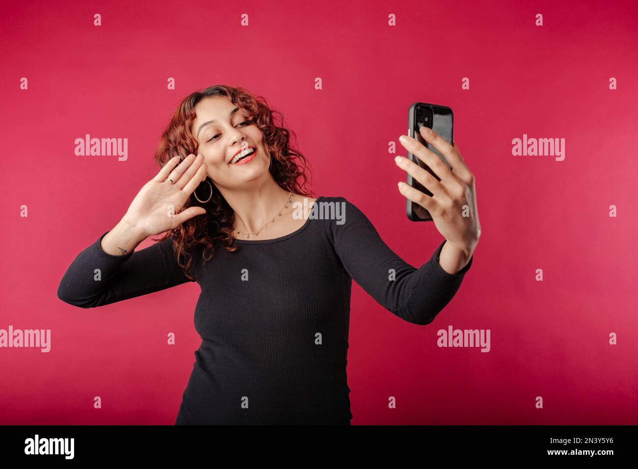 Bella donna rossa indossando abito nero in piedi isolato su sfondo rosso utilizzando lo smartphone per videochiamare. Chat video, parlare sul cellulare Foto Stock