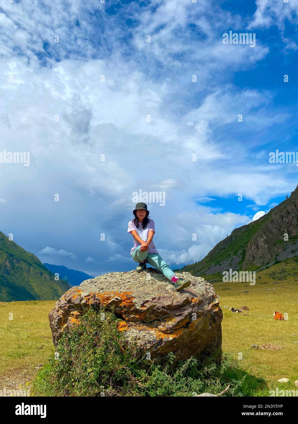 Una ragazza viaggiatore asiatica in un cappello e occhiali si siede con la gamba distesa su uno sfondo di pietra delle montagne di Altai. Telaio verticale. Foto Stock