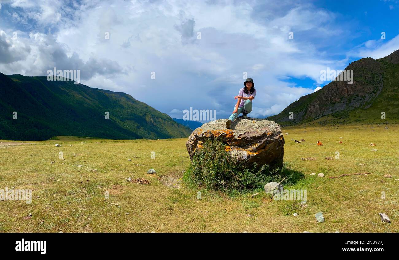 Una ragazza viaggiatore asiatica in un cappello e occhiali si siede con la gamba distesa su uno sfondo di pietra delle montagne di Altai. Foto Stock