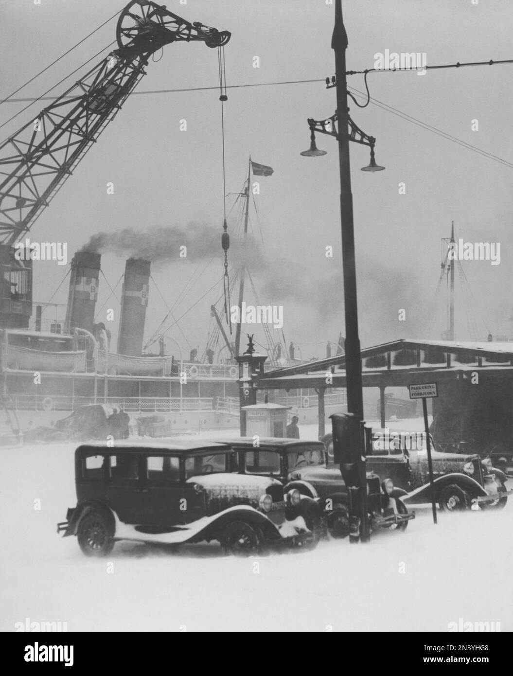 Inverno negli anni '1920s. Una giornata invernale innevata e ventosa a Stoccolma. Tre automobili sono viste parcheggiate davanti alla banchina. Una nave a vapore con fumo proveniente dal suo camino con una gru che carica o scarica qualcosa Svezia 1928 Foto Stock
