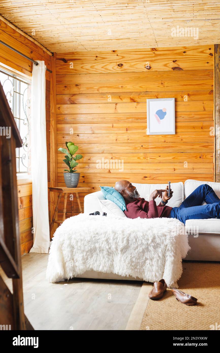 Uomo anziano afro-americano che usava lo smartphone mentre era sdraiato sul divano in una cabina di tronchi Foto Stock