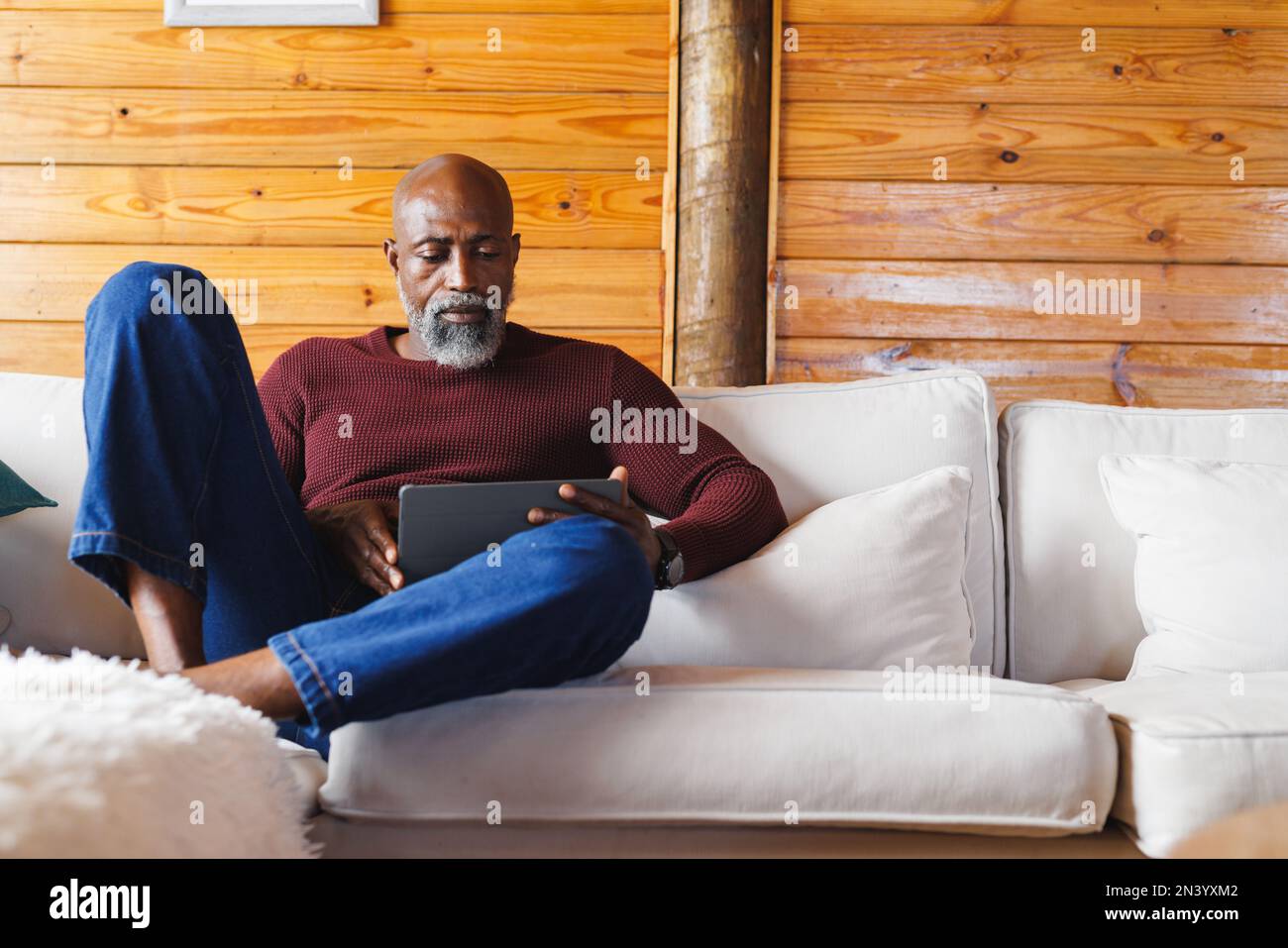 Uomo anziano afro-americano calvo che utilizza un tablet digitale mentre si siede sul divano in una cabina di tronchi Foto Stock