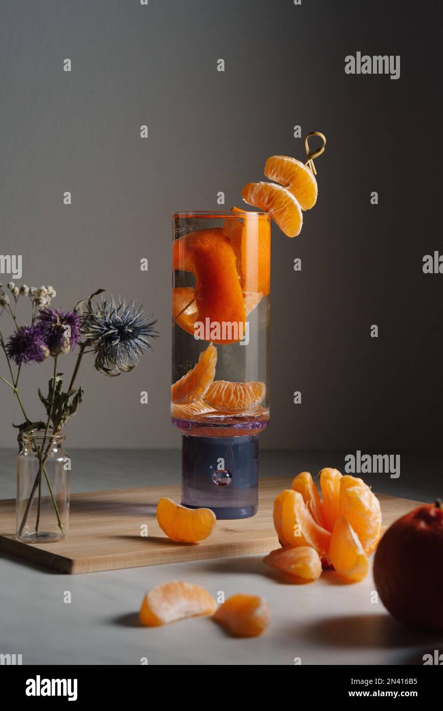 un bicchiere d'acqua viola e la pelle di clementina in esso, un po' di frutta fresca, fiori secchi, clementine e uno sfondo grigio Foto Stock un bicchiere d'acqua viola e la pelle di clementina in esso, un po' di frutta fresca, fiori secchi, clementine e uno sfondo grigio Foto Stock