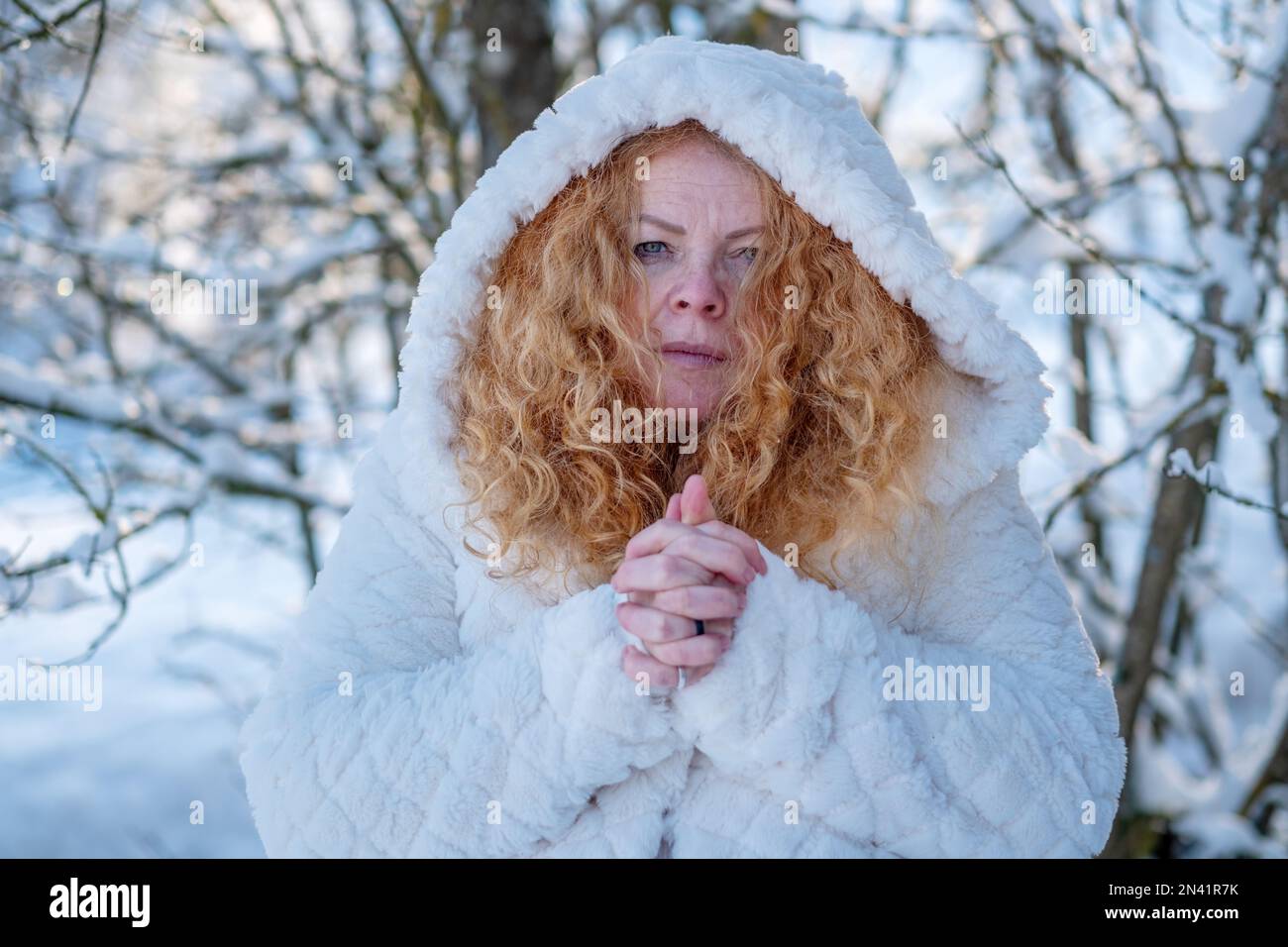 ritratto di bella donna sexy rossa matura, con capelli ricci rossi in cappotto bianco con cappuccio in inverno natura controluce sole, albero nevoso, copia spazio Foto Stock