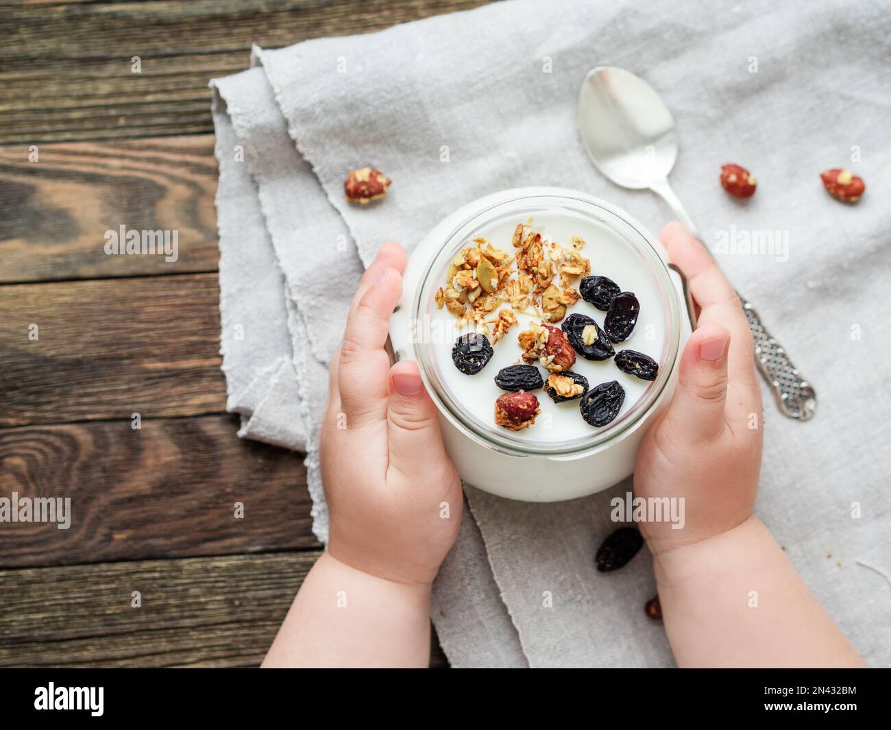Yogurt naturale fatto in casa in un vasetto di vetro. Cibo sano per la colazione nelle mani dei bambini. Vasetto con muesli su tovaglia di lino su tovaglia di legno Foto Stock