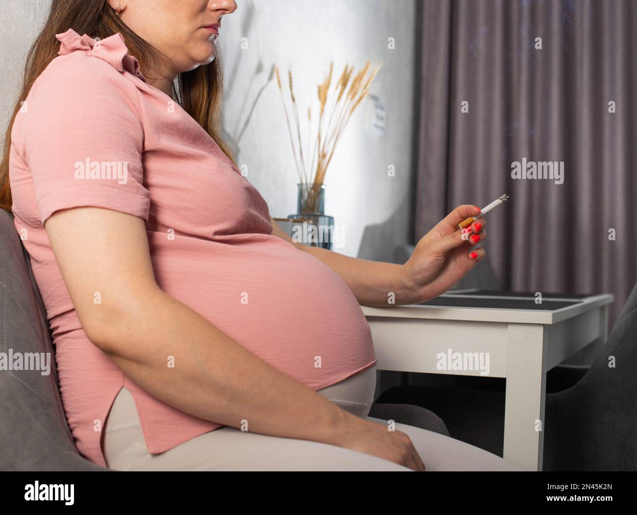 Una ragazza incinta con una grande pancia siede in cucina al tavolo e fuma. Fumo e gravidanza. Danno alla salute del bambino e della madre, tobac Foto Stock