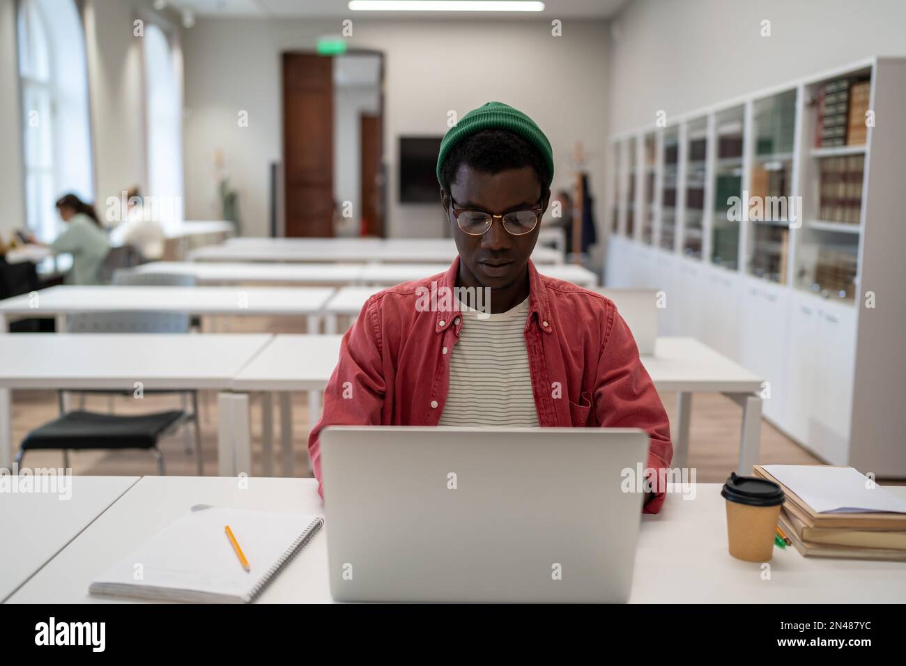 Focalizzato afro americano uomo in occhiali di studio on-line sul computer portatile, mentre siede in biblioteca universitaria Foto Stock