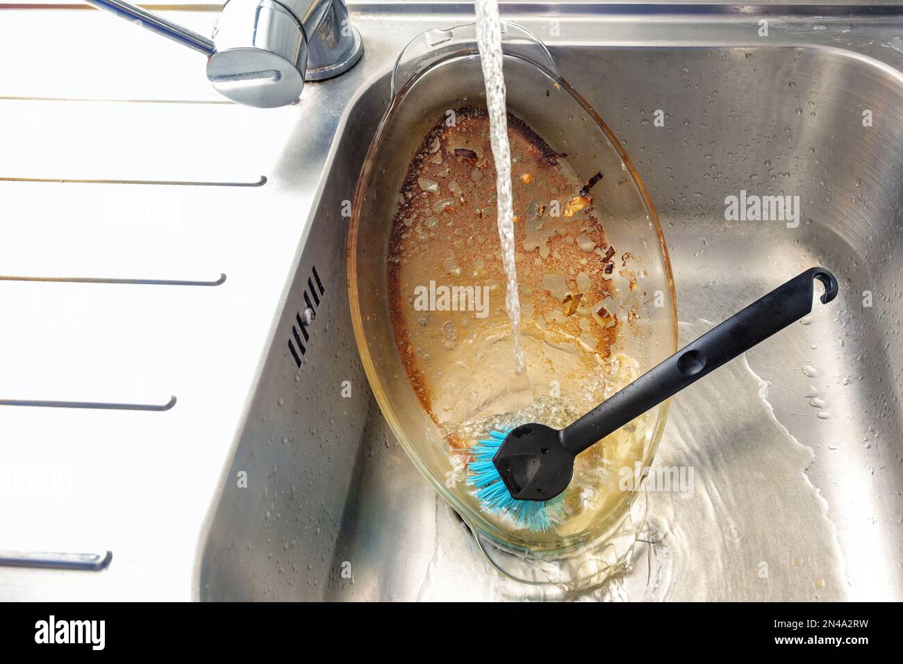 La casseruola di vetro sporca con grasso bruciato viene pulita nel lavello sotto un getto d'acqua con un pennello, il lavaggio dei piatti a mano, noioso chore domestico, copia Foto Stock