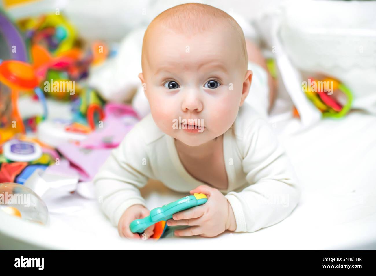 Un bambino divertente in vestiti bianchi si trova su un lettino vicino ai giocattoli. Una bambina cute poco neonato si trova su un letto tra giocattoli colorati per i bambini. Il primo educativo Foto Stock
