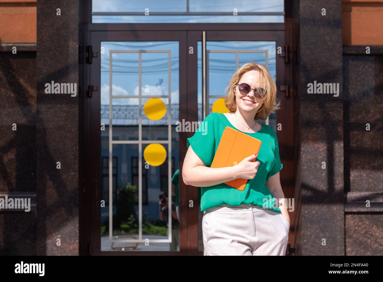 Una ragazza studentesca felice che sorride negli occhiali da sole, tenendo un tablet digitale sullo sfondo dell'edificio universitario. Una ragazza attraente che guarda ca Foto Stock