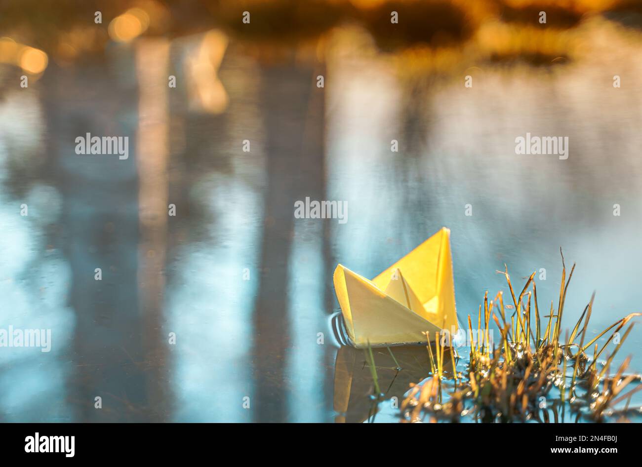 Imbarcazione di carta galleggiante gialla. Nave a vela colorata in grande pozza di primavera blu, acqua del fiume su strada invernale. Caldo tempo piovoso bagnato, erba vecchia. Ciao sp Foto Stock