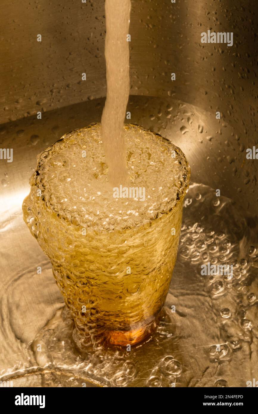 L'acqua che fluisce da un rubinetto riempie un bicchiere a traboccante. Formato verticale. Foto Stock