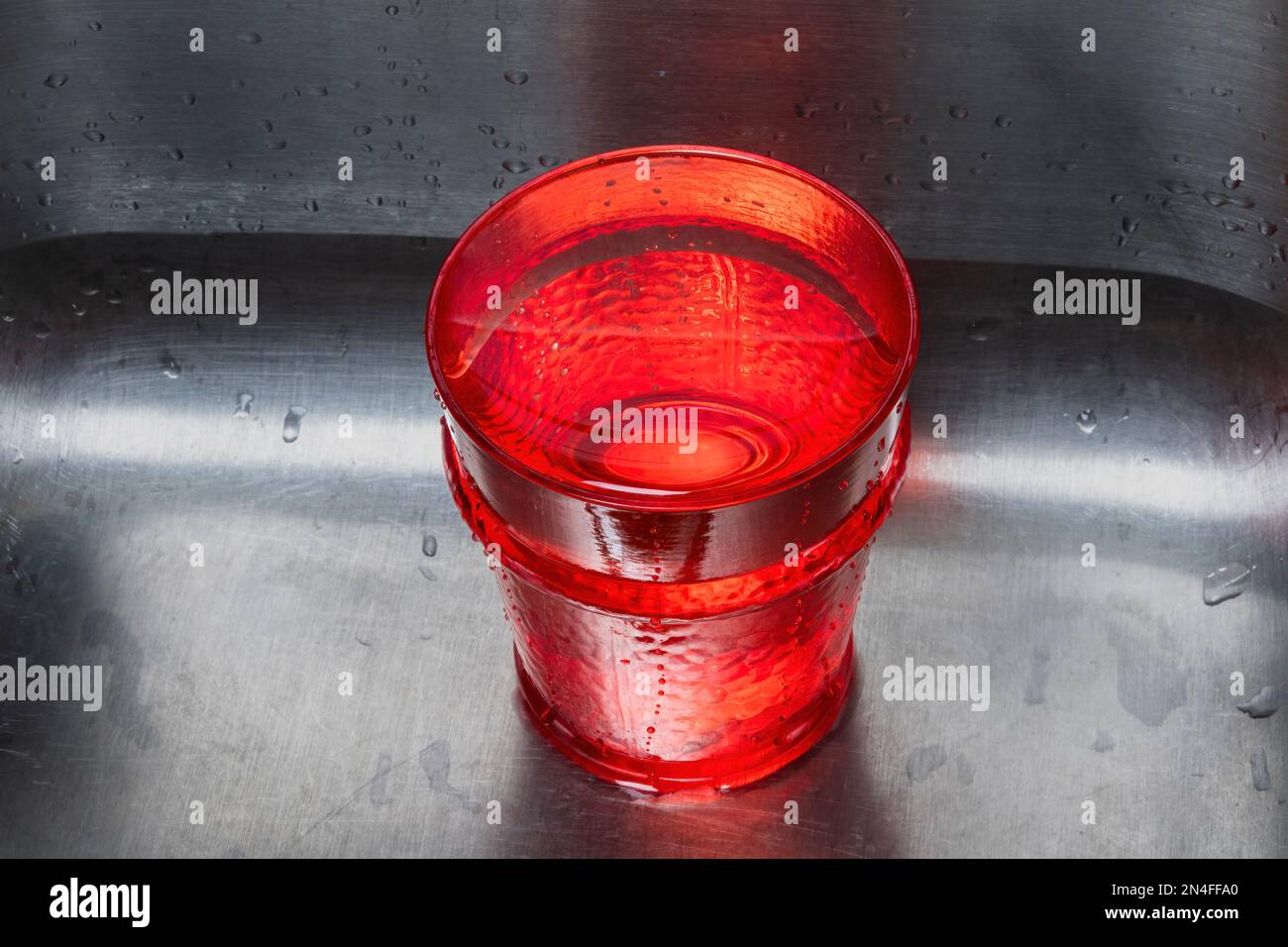 Una tazza da bere rossa piena d'acqua in un lavandino in acciaio inossidabile. Invertire lo spazio di copia su entrambi i lati. Foto Stock