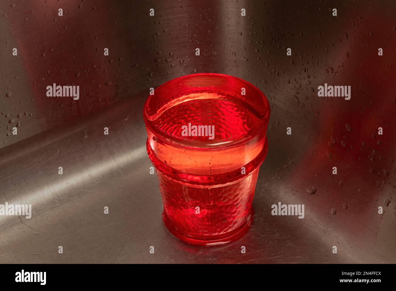 Vetro rosso pieno d'acqua in un angolo del lavello in acciaio inossidabile. Invertire tutto lo spazio di copia. Foto Stock