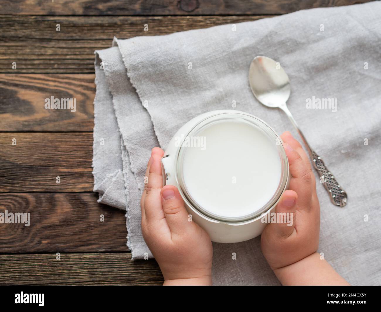 Yogurt naturale fatto in casa in un vasetto di vetro. Cibo sano per la colazione nelle mani dei bambini. Vasetto con crema acida su tovaglia di lino su legno Foto Stock