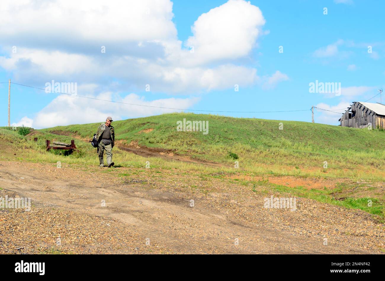 Sorridente uomo di villaggio Asian Yakut turista giù per la collina sulla strada con uno zaino sullo sfondo di erba e nuvole. Foto Stock