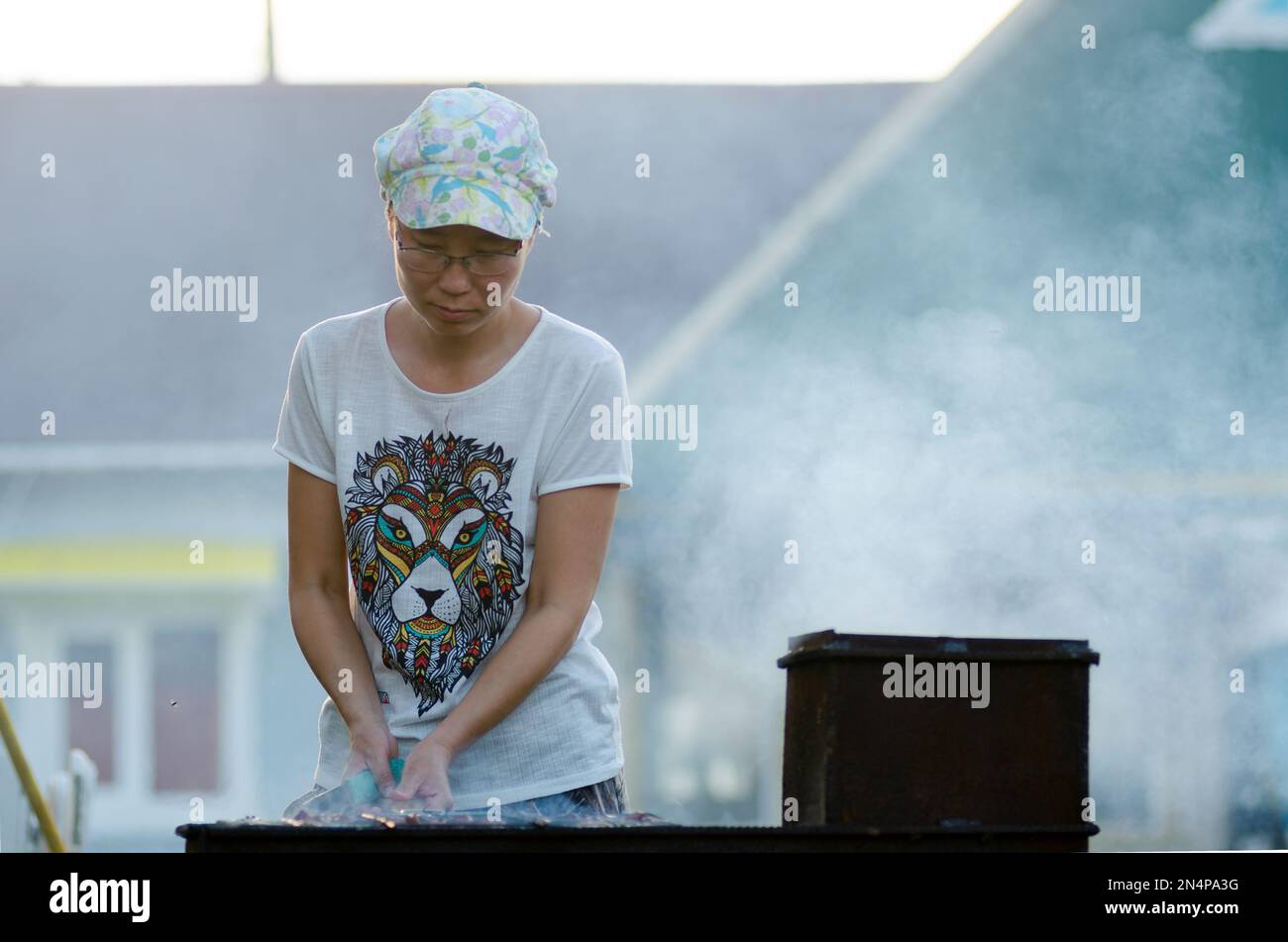 Yakut ragazza asiatica con bicchieri e un tappo prepara barbecue a una grande griglia nel fumo sullo sfondo di una casa residenziale privata nel nord Foto Stock