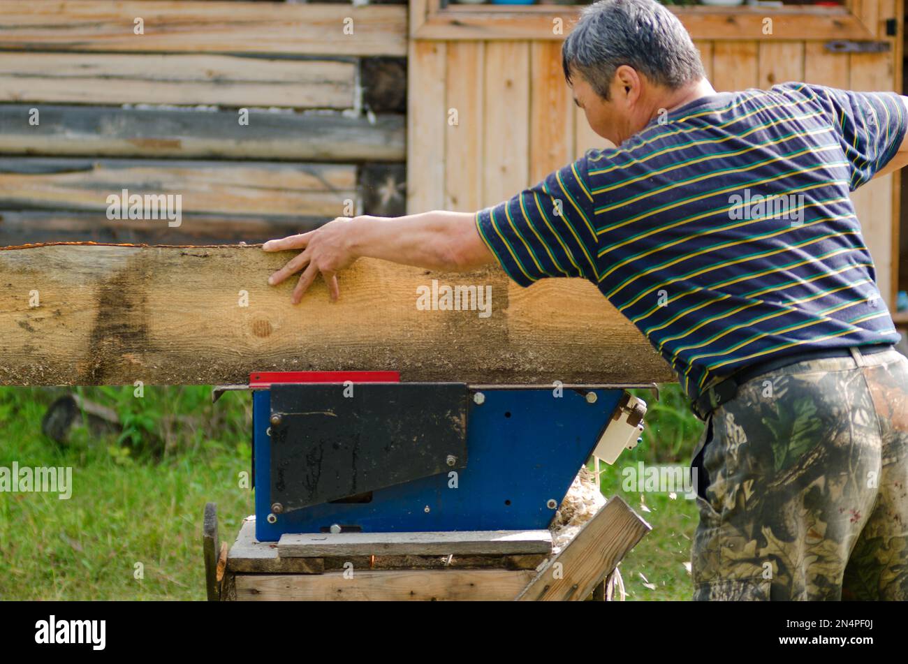 L'uomo Yakut adulto sega un bordo su una sega contro una casa di legno nel villaggio di Yakut. Foto Stock