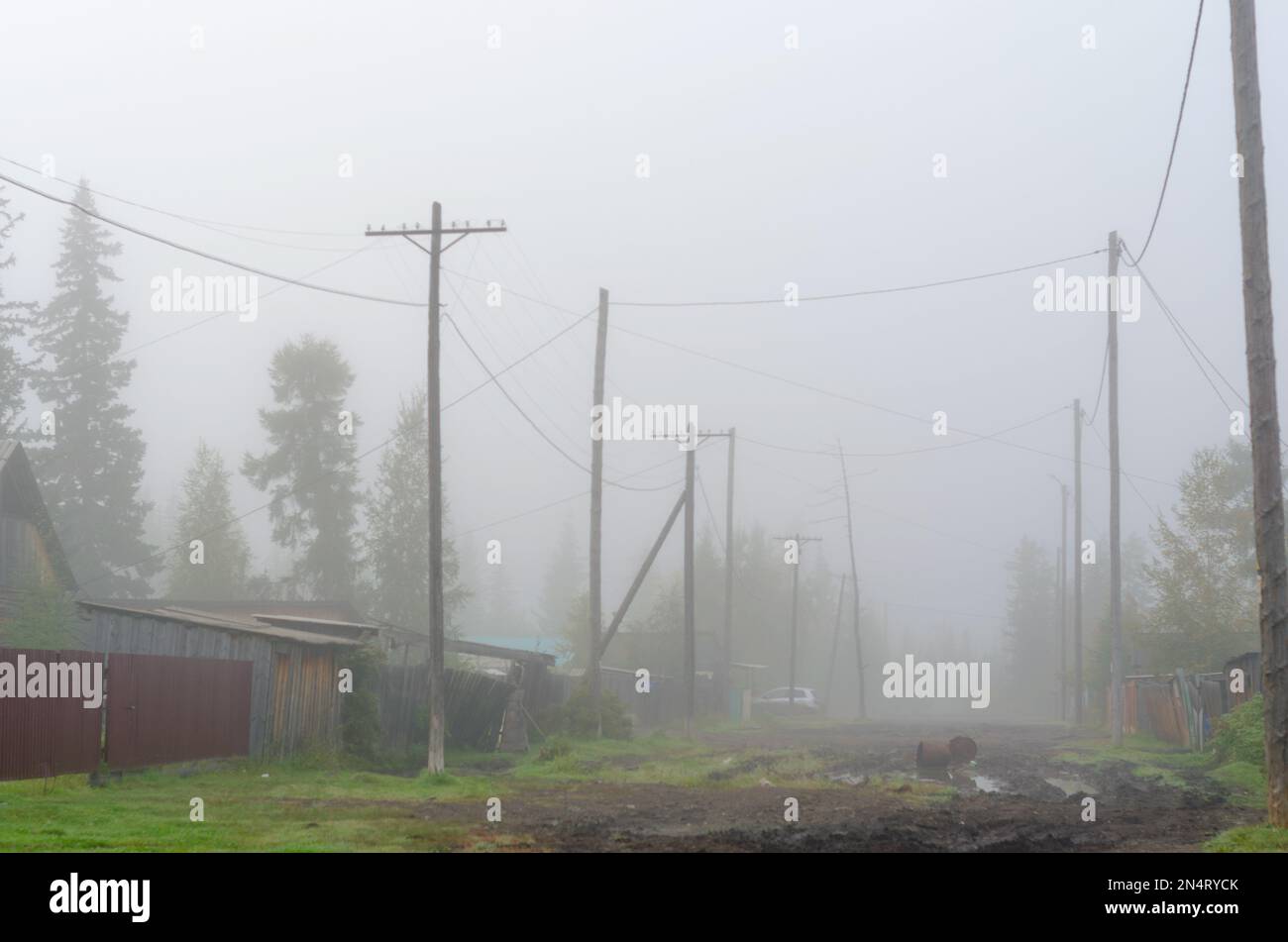 La strada del villaggio di Yakut nord di ulus suntar nella nebbia mattutina con pozzanghere sulla strada e auto in piedi dietro i poli della linea di alimentazione e w Foto Stock