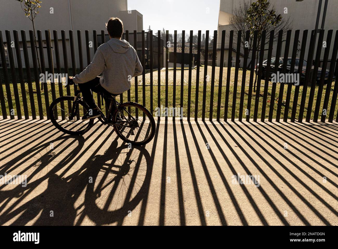 ragazzo in bicicletta passa accanto a un marciapiede pavimentato con ombra a strisce dalla recinzione Foto Stock