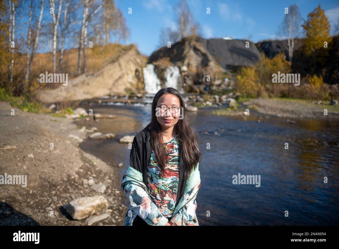 Yakut ragazza asiatica in occhiali che si posa sorridendo sullo sfondo di una piccola cascata in autunno. Foto Stock