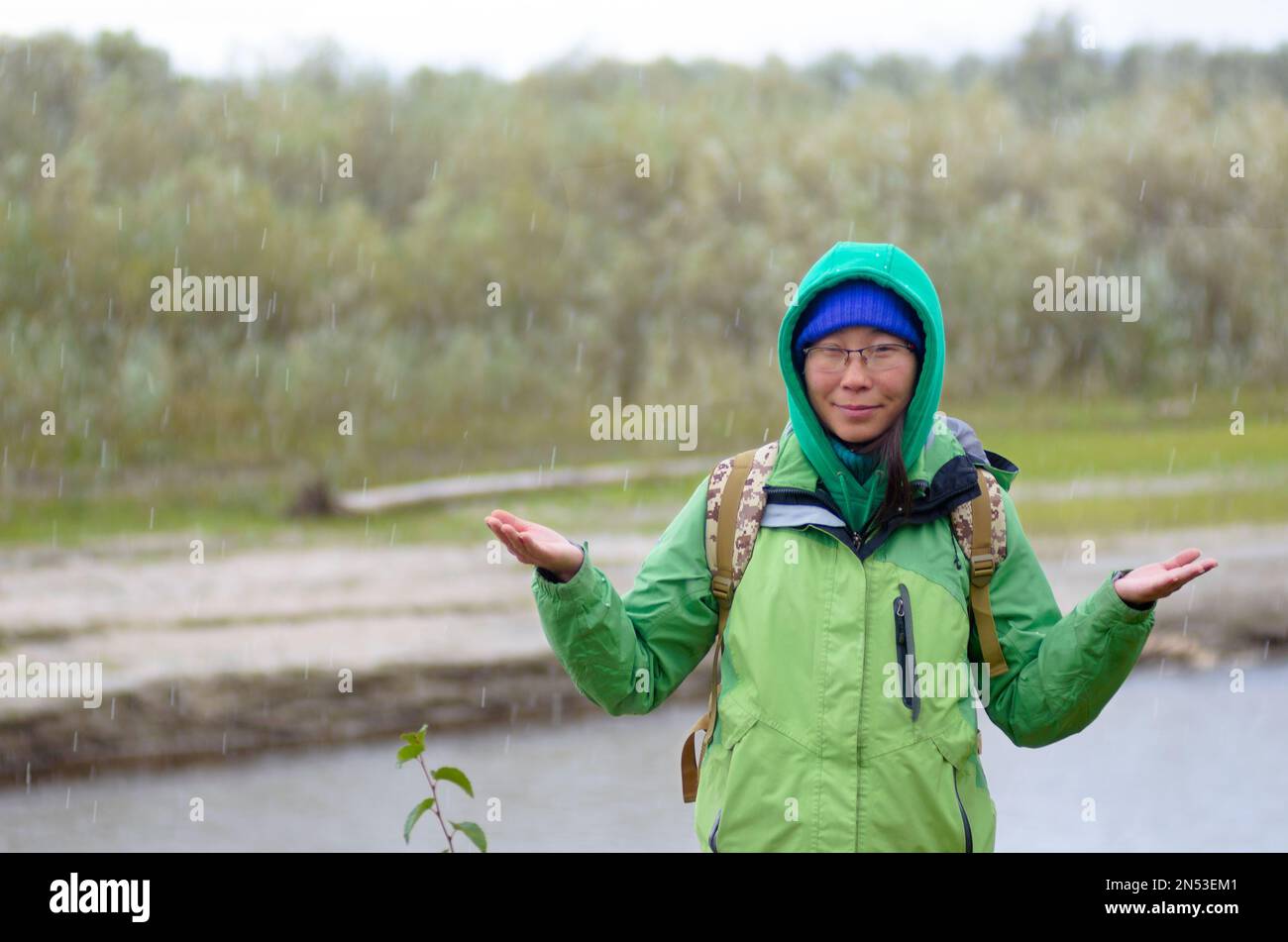 Yakut ragazza asiatica turista in una giacca cappello e cappuccio e con uno zaino è sorridente, grandine e pioggia tenendo le mani contro lo sfondo della riv Foto Stock