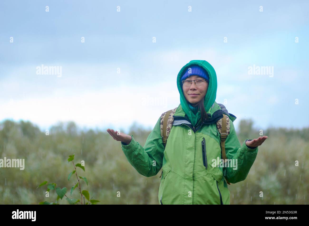Yakut ragazza asiatica turista in una giacca cappello, cappuccio e occhiali, con uno zaino è sorridente, grandine, pioggia e neve tenendo le mani contro lo zaino Foto Stock
