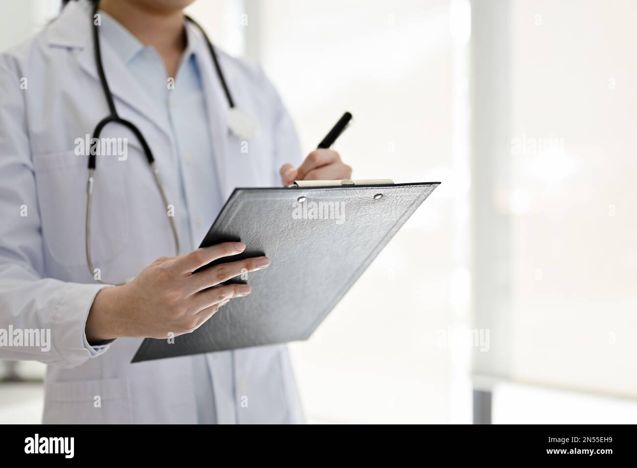 Immagine ritagliata di un medico femminile asiatico professionista in abito bianco in piedi nella sala esame, scrivendo qualcosa su una clipboard medica, o recor Foto Stock