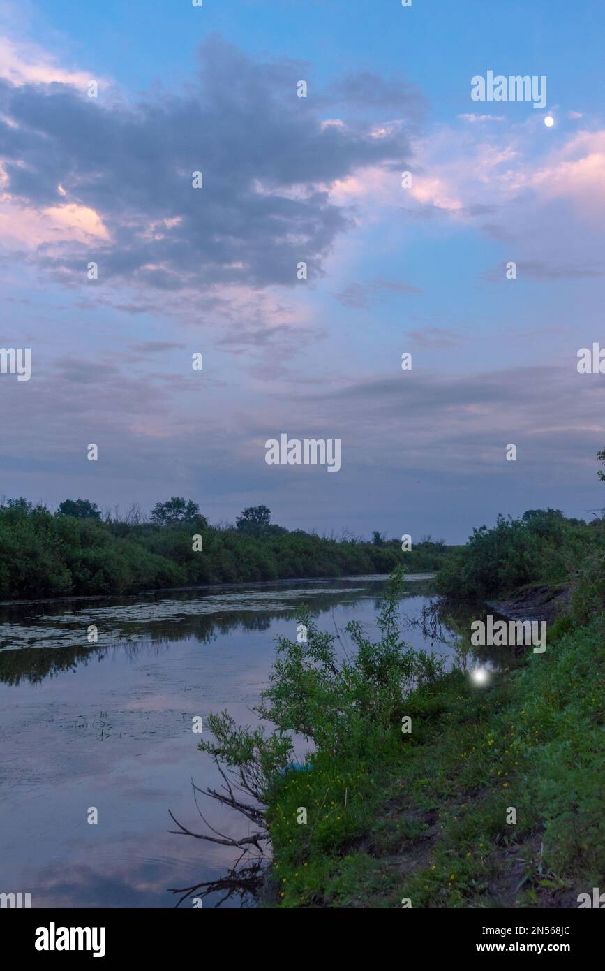 Il riflesso della luna e delle nuvole cielo tramonto al crepuscolo nelle acque del fiume vicino alle rive con erba verde e alberi. Foto Stock