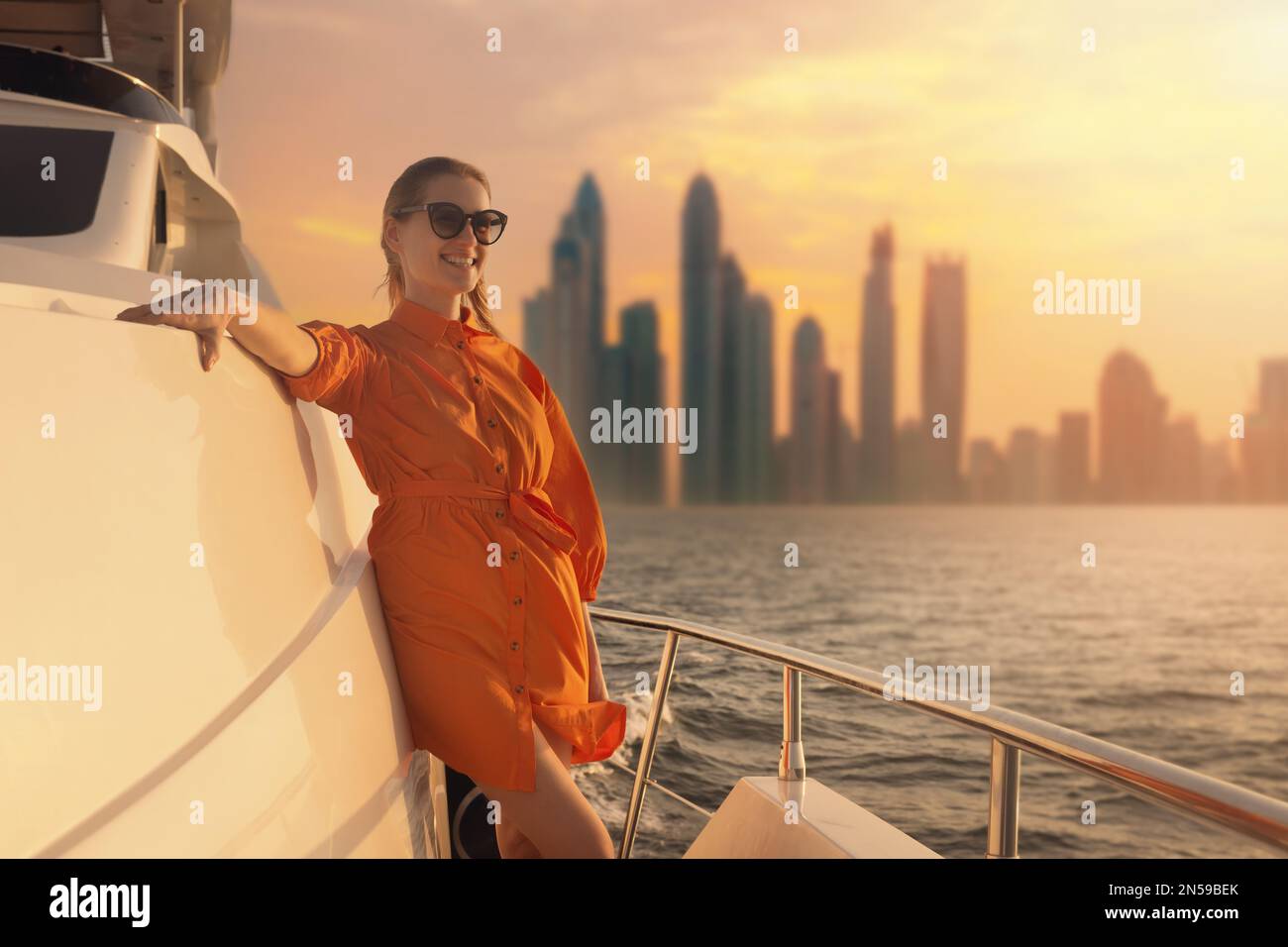 Donna in abito arancione sul ponte di yacht di lusso con skyline di Dubai sullo sfondo al tramonto. spazio di copia Foto Stock