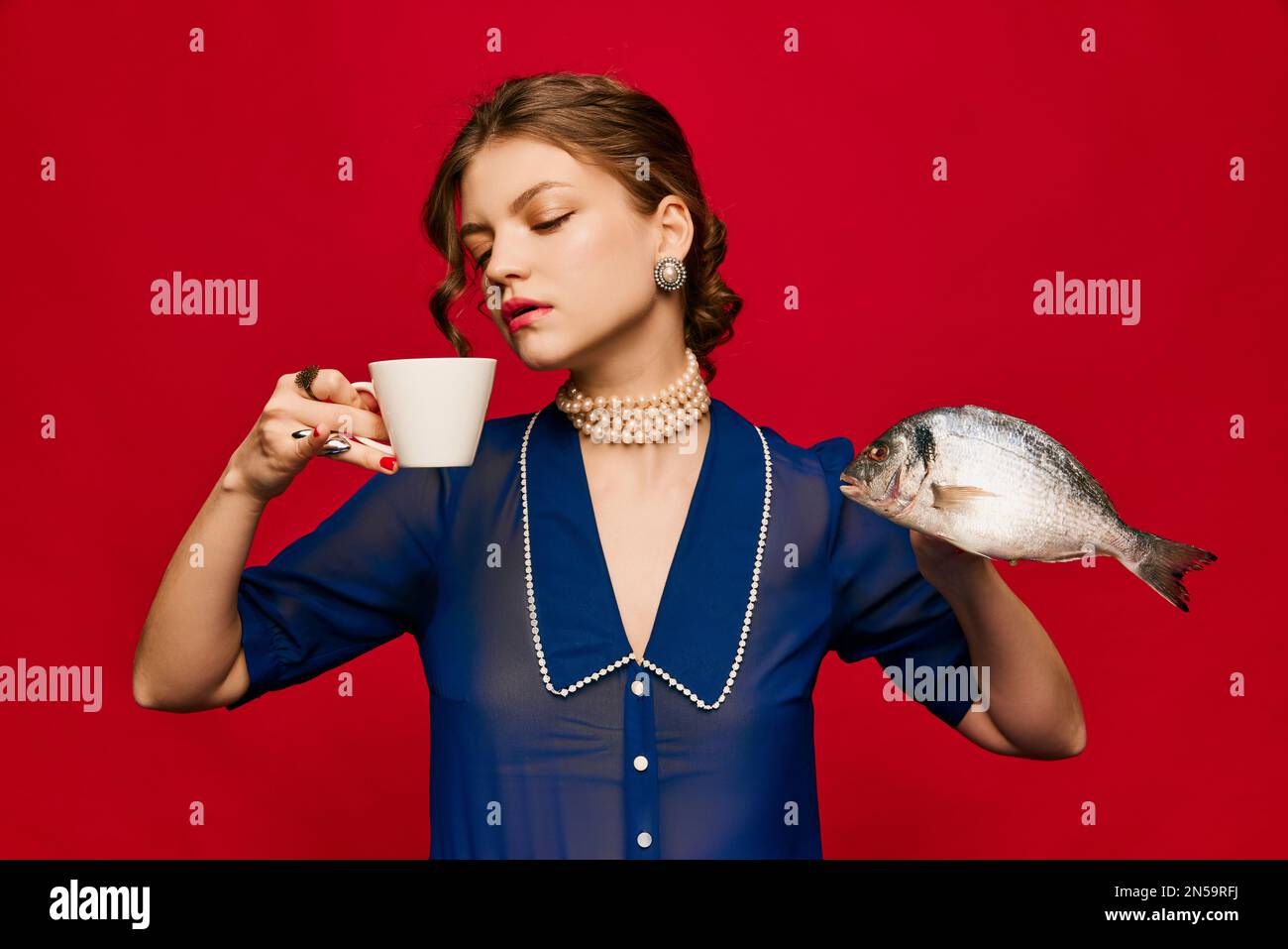 Gusto delizioso e strano. Giovane donna stravagante con tazza di caffè e pesce crudo su sfondo rosso brillante. Vintage, stile retrò. Colori complementari Foto Stock