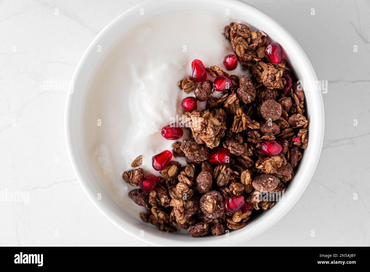 Ciotola di yogurt greco con granola di avena al cioccolato e semi di melograno su tavola di marmo bianco. Vista dall'alto. Colazione sana Foto Stock