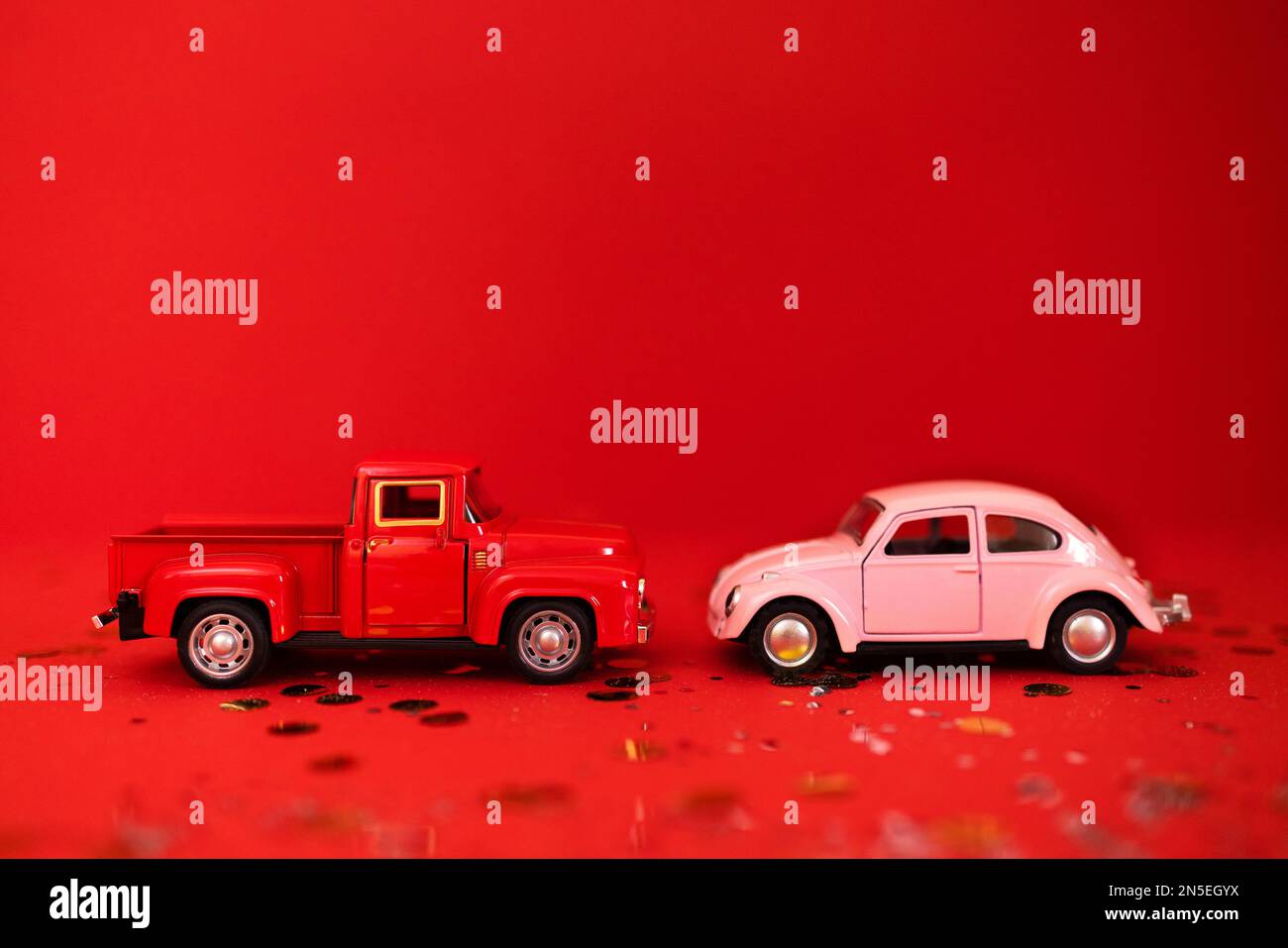 Due macchinine su sfondo rosso con luccichio e coriandoli. Un autocarro rosso e una macchina rosa per passeggeri si trovano uno di fronte all'altro. San Valentino concep Foto Stock