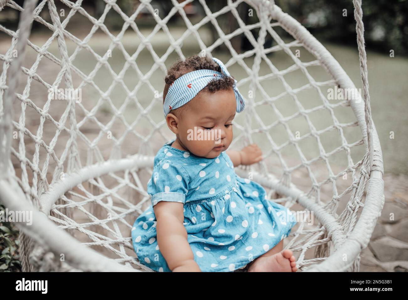 La bambina swarthy in vestito blu del punto della polka ha emozione positiva seduta sul Cocoon bianco della corda del macramè del cotone appeso. Bambino africano americano rel Foto Stock