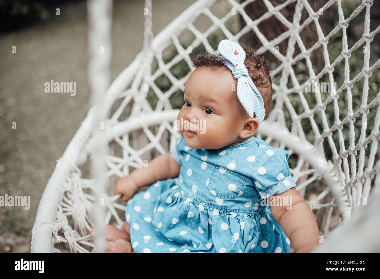 La bambina swarthy in vestito blu del punto della polka ha emozione positiva seduta sul Cocoon bianco della corda del macramè del cotone appeso. Bambino africano americano rel Foto Stock